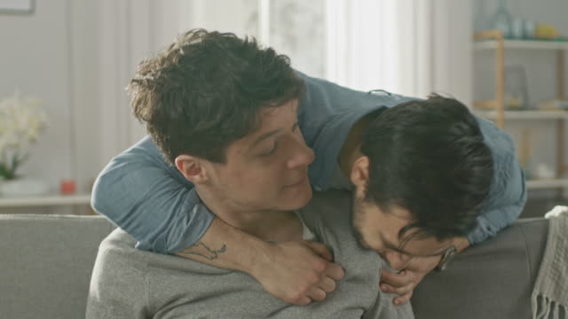 Cute-Male-Queer-Couple-Spend-Time-at-Home.-Young-Man-Uses-a-Laptop,-His-Partner-Comes-From-Behind-and-Gently-Embraces-Him.-They-Laugh-and-Touch-Hands.-Room-Has-Modern-Interior.