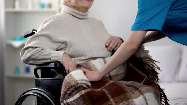 Enfermera-cubriendo-suavemente-con-mujer-de-manta-en-silla-de-ruedas,-cuidado-de-ancianos
