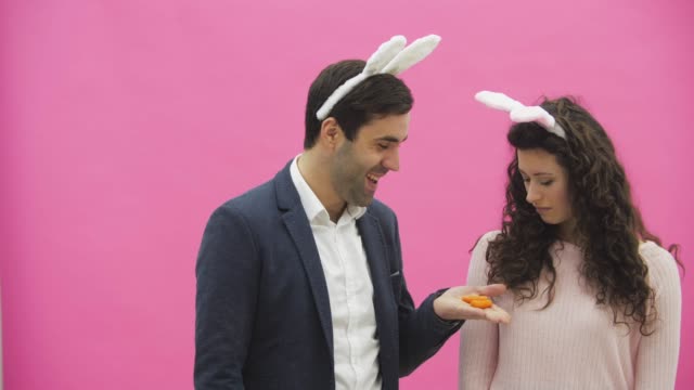 Young-couple-are-beautiful-on-pink-background.-During-this-time,-they-are-dressed-in-rabble-ears.-Looking-at-each-other.-A-man-shows-carrots,-his-wife-does-not-like-the-size-of-carrots-and-she-throws-out.