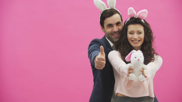 Young-creative-couple-on-pink-background.-With-hackneyed-ears-on-the-head.-During-this,-a-wife-holds-a-soft-toy-hare,-a-man-shows-a-class.-Giving-a-genuine-smile.