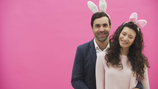 Young-creative-couple-on-pink-background.-With-hackneyed-ears-on-the-head.-During-this,-two-show-the-gestures-of-the-class-and-look-at-one-another-together.