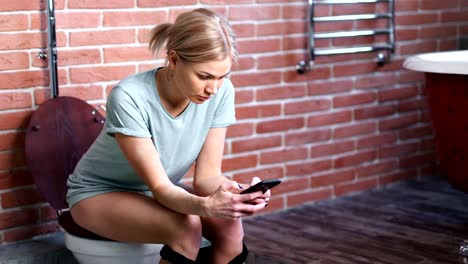 Confident-young-woman-sitting-on-toilet-in-bathroom-using-smartphone-medium-long-shot