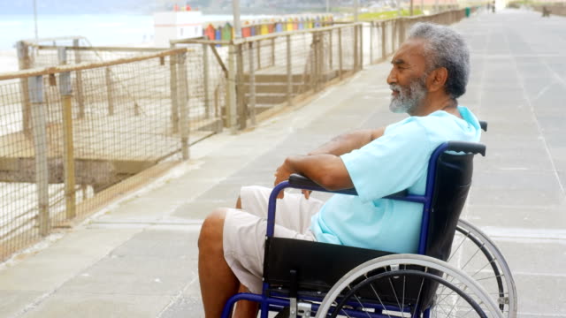 Seitenansicht-von-nachdenklich-behinderten,-aktiven,-ranghohen-Afroamerikaners-im-Rollstuhl-auf-der-Promenade-4k