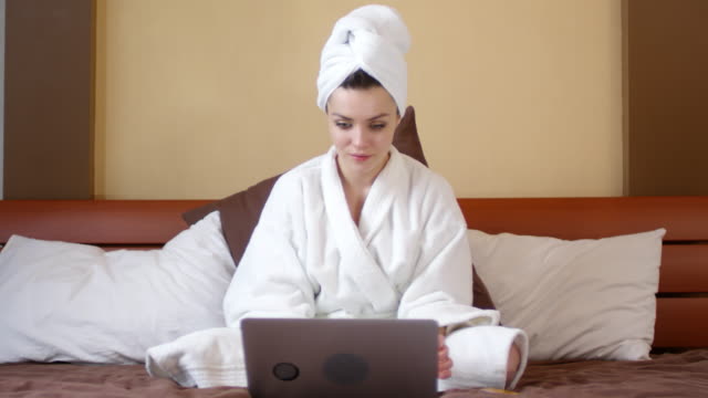 Fröhliche-Frau-in-Bathrobe-sitzt-auf-Bett-und-benutzt-Laptop