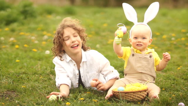 Two-brothers,-a-teenager-and-a-baby-are-resting-in-a-park-on-a-glade.-Easter-concept,-bunny-ears,-easter-basket-and-eggs.-A-dog-runs-past-them