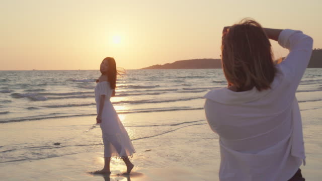Young-Asian-lesbian-couple-using-camera-taking-photo-each-other-near-beach.-Beautiful-women-lgbt-couple-happy-romantic-moment-when-sunset-in-evening.-Lifestyle-lesbian-couple-travel-on-beach-concept.