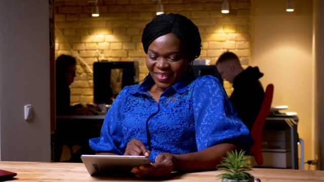 Closeup-shoot-of-happy-adult-african-american-businesswoman-using-the-tablet-and-laughing-cheerfully-in-the-office-indoors