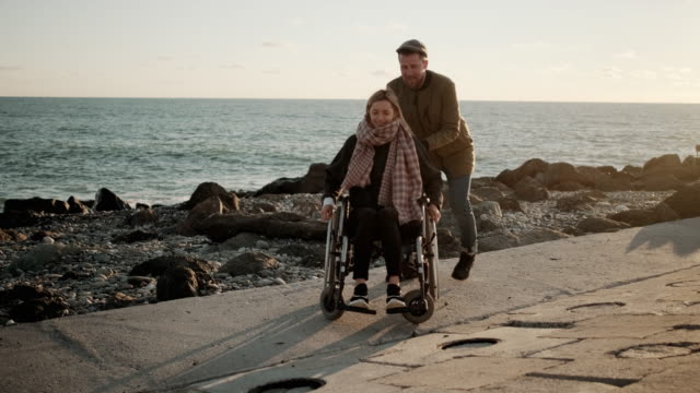 Loving-man-is-whirling-wheelchair-with-young-woman-on-sea-coast,-rejoicing