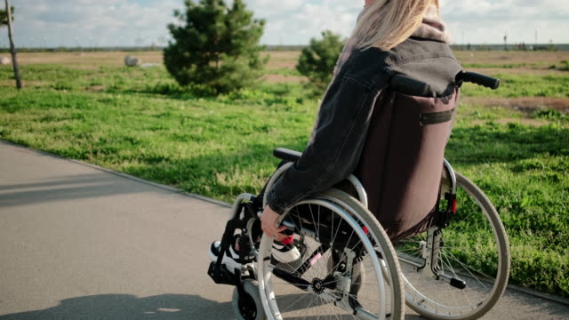 Young-woman-is-moving-by-road-in-park-riding-invalid-chair-in-sunny-weather