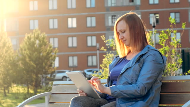 Woman-using-tablet-computer-sitting-on-bench-in-city