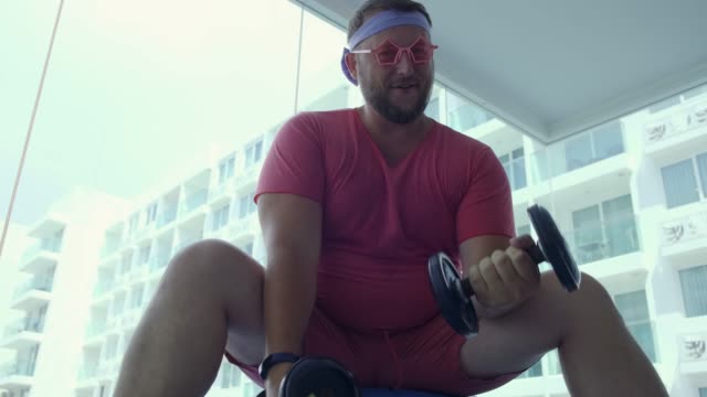 Funny-fat-male-in-pink-glasses-and-in-a-pink-t-shirt-is-engaged-with-dumbbells-on-a-fit-ball-in-the-gym-depicting-a-girl