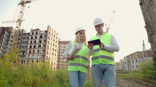 Business,-building,-industry,-technology-and-people-concept---smiling-builder-in-hardhat-with-tablet-pc-computer-along-with-woman-with-drawings-of-builders-at-construction-site.