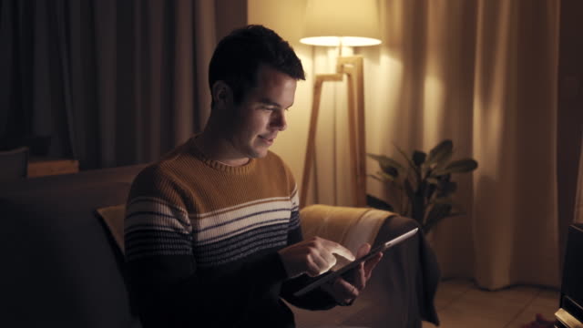 Glücklicher-junger-Mann-sitzt-auf-dem-Sofa-beschäftigt-mit-digitalen-Tablet-halten-in-den-Händen-in-der-späten-Nacht