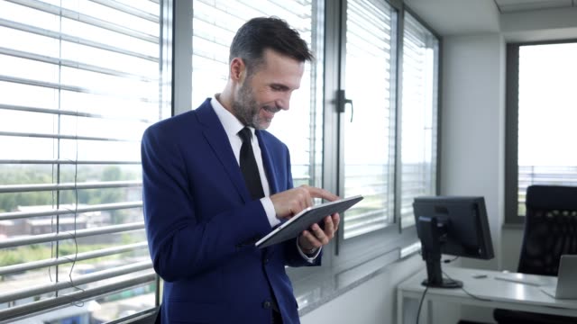 Hombre-de-negocios-sonriente-usando-tableta-y-apoyado-en-la-ventana-de-la-oficina