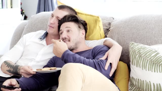 Schwules-Paar-entspannend-auf-der-Couch.-Fernsehen-und-Kekse-genießen.