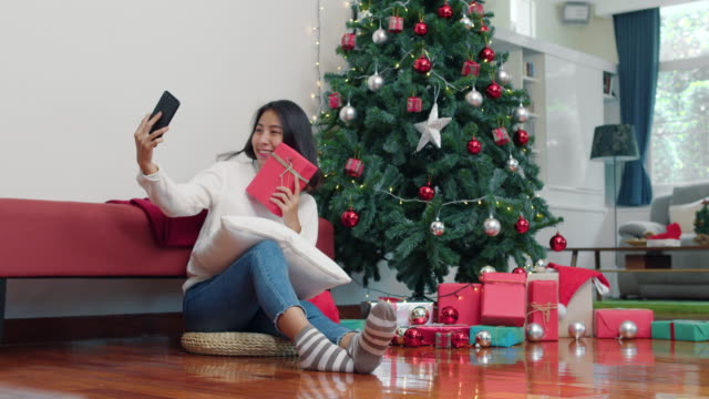 Asian-women-celebrate-Christmas-festival.-Female-teen-relax-happy-holding-Gift-and-using-smartphone-selfie-with-Christmas-tree-enjoy-xmas-winter-holidays-in-living-room-at-home.
