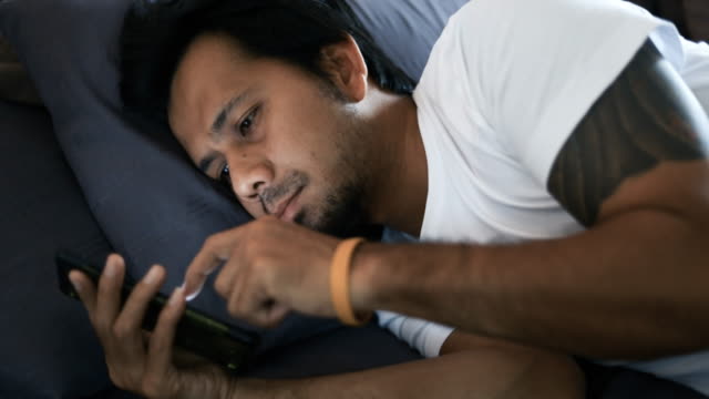 junger-asiatischer-Mann-mit-seinem-Smartphone-am-Abend,-während-er-im-Bett-vor-dem-Schlafengehen-liegt