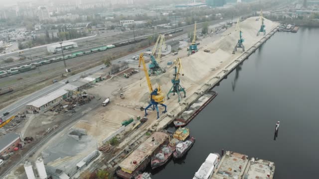 Industrial-city-part-with-river-port-and-docks-with-yellow-construction-and-cargo-cranes-extracting-sand-and-rocks-from-barge