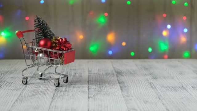 Hand-drücken-Mini-Einkaufswagen-mit-Weihnachtsbaum-und-Geschenke-auf-dem-Hintergrund-der-LED-Lampen-Girlande.