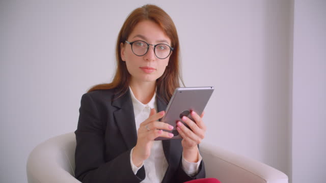Nahaufnahme-Porträt-der-jungen-kaukasischen-Geschäftsfrau-in-Brille-SMS-auf-dem-Tablet-aussehenta-Kamera-lächelnd-glücklich-sitzend-im-Sessel