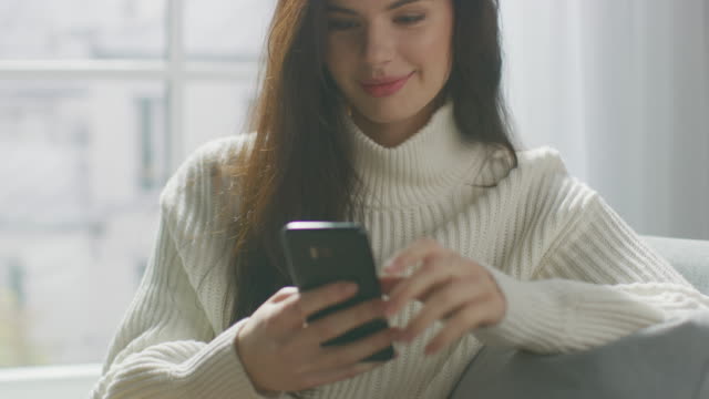 Beautiful-Young-Woman-Using-Smartphone-Smilingly,-while-Sitting-on-the-Chair.-Sensual-Girl-Wearing-Sweater,-Surfs-Internet,-Posts-on-Social-Media-while-Relaxing-in-Cozy-Apartment.-Closeup-Portrait