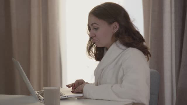 Close-up-of-positive-Caucasian-girl-using-laptop-in-the-morning.-Young-beautiful-woman-in-white-bathrobe-using-social-media.-Internet,-online,-lifestyle,-leisure.