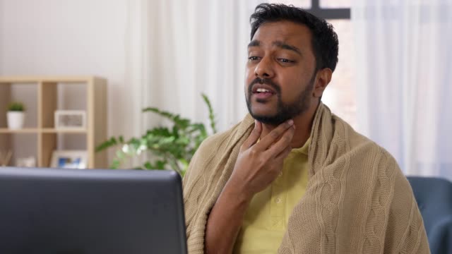 healthcare,-technology-and-people-concept---sick-indian-man-in-blanket-with-sore-throat-having-video-call-on-laptop-computer-at-home