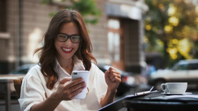 Business-female-in-glasses,-white-shirt.-Sitting-at-table-in-outdoor-cafe.-Looking-at-contract-on-paper,-then-at-smartphone-and-rejoicing.-Slow-motion