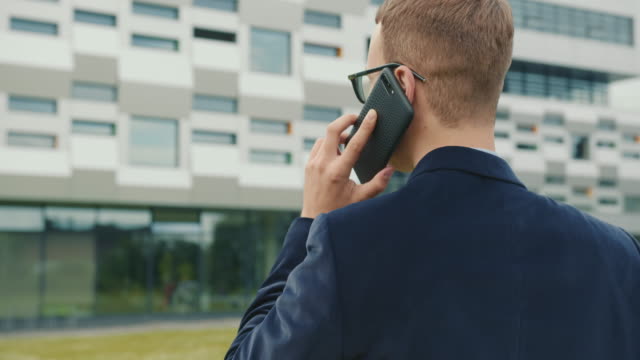 The-businessman-is-going-and-talking-on-the-phone.-He's-wearing-a-suit-and-glasses.-The-business-center-is-in-the-background.-Close-up-shooting.-4K