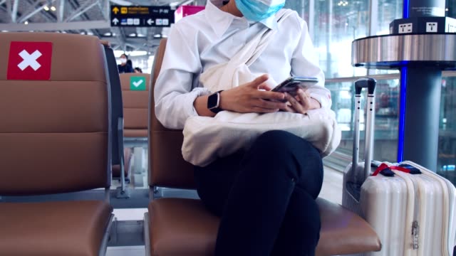 Empresaria-que-usa-protección-de-máscara-quirúrgica-y-utiliza-teléfonos-inteligentes-de-verificación-de-los-medios-sociales-mientras-está-sentado-una-silla-en-la-terminal-del-aeropuerto-y-mantener-una-distancia-social-durante-la-pandemia-COVID-19.
