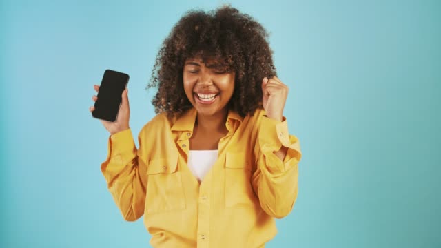Afro-american-lady-looking-at-smartphone,-saying-yes,-showing-you-the-screen-and-smiling-posing-on-blue-background