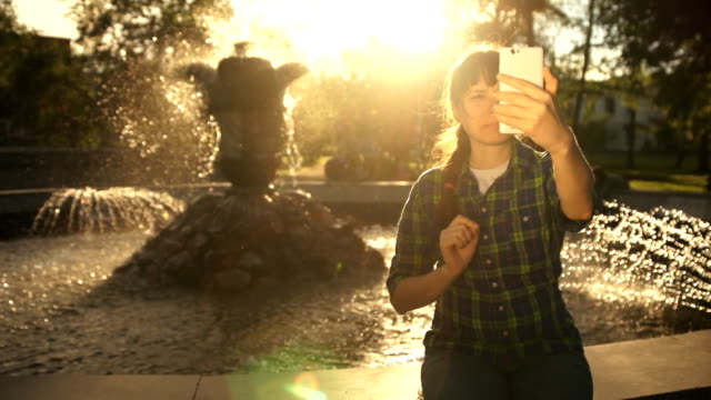 Eine-junge-Frau-nimmt-ein-Selbstporträt-auf-ein-Telefon-in-der-Nähe-eines-Brunnens-in-einem-öffentlichen-park