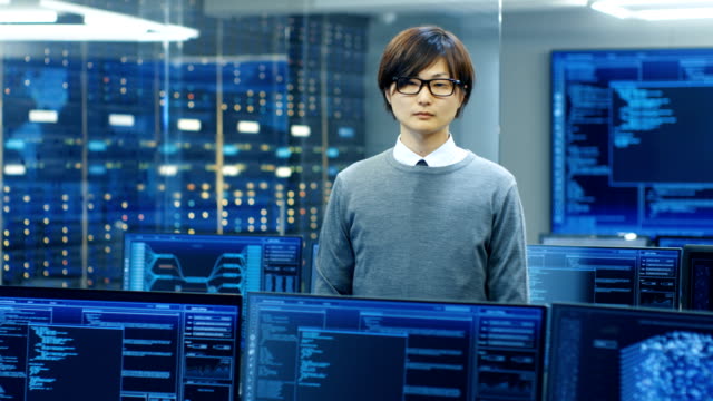 In-the-System-Control-Room-Technical-Operator-Stands-and-Looks-at-the-Camera.-He's-Surrounded-by-Multiple-Displays-Showing-Graphics.-IT-Technician-Works-on-Artificial-Intelligence,-Big-Data-Mining,-Neural-Network-Project.
