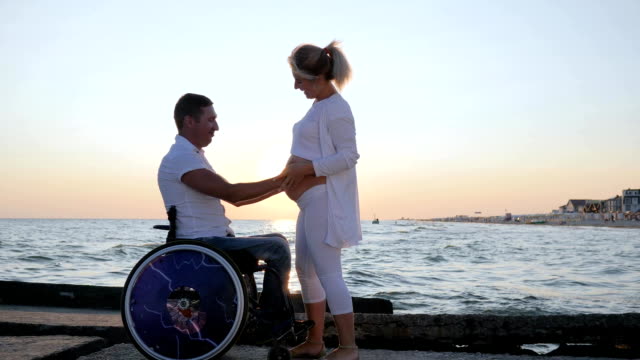 Behinderten-im-Rollstuhl-hört-Bauch-seiner-Frau-am-Meer-Hintergrund