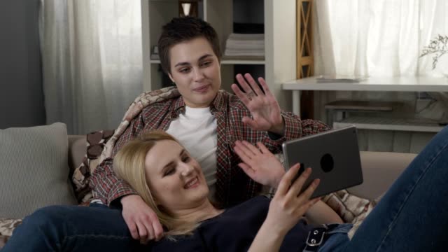 Lesbian-couple-is-resting-on-the-couch,-and-having-video-conversation-with-friend,-waving-their-arms,-video-call-60-fps
