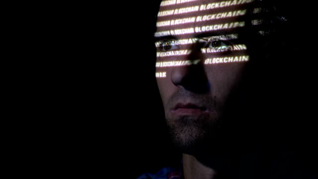 Nahaufnahme-eines-erwachsenen-Mannes-mit-Bart-Blick-auf-einem-Bildschirm,-während-das-Blockchain-Symbol-auf-seinem-Gesicht-reflektiert-wird