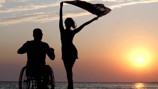 shapely-girl-is-standing-on-jetty-and-waving-cloth-in-front-of-man-disabled-into-wheel-chair-on-background-of-sunset-at-summer-weekend