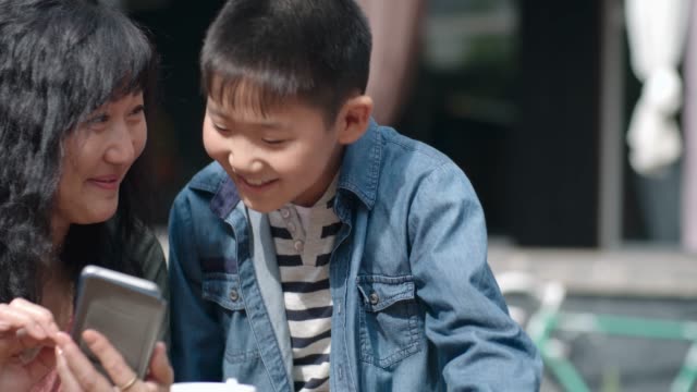 Mujer-asiática-y-niño-sonriendo-y-usando-el-teléfono-inteligente-al-aire-libre