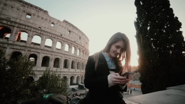 Young-attractive-woman-standing-near-the-Colosseum-in-Rome,-Italy-and-using-the-smartphone.-Smiling-girl-chatting