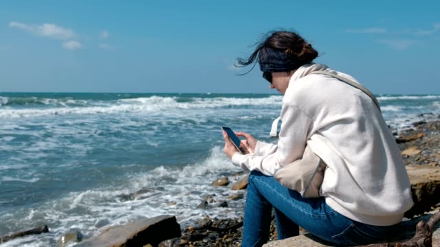 Mujer-juega-en-teléfono-sentado-en-la-orilla-del-mar-en-otoño.