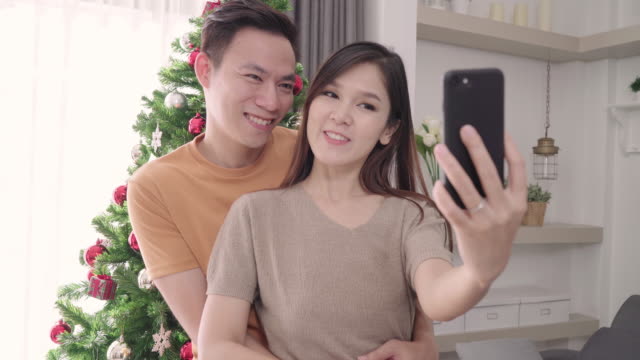 Asian-paar-mit-Smartphone-Selfie-mit-Weihnachtsbaum-schmücken-ihre-Wohnzimmer-zu-Hause-in-Christmas-Festival.-Lifestyle-Frau-und-Mann-glücklich-feiern-Sommer-Weihnachten-und-Neujahr-Konzept.