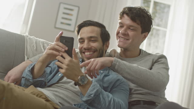 Süße-männliche-Gay-paar-verbringen-Zeit-zu-Hause.-Sie-sind-unten-auf-dem-Sofa-liegen-und-ein-Smartphone-benutzen.-Stöbern-sie-Online.-Partners-Hand-ist-um-seine-Geliebte.-Sie-Lächeln-und-lachen.-Zimmer-ist-Modern-eingerichtet.