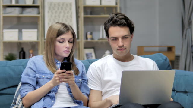 Junge-Paar-Gespräch-während-mittels-Smartphone-und-Laptop-zu-Hause
