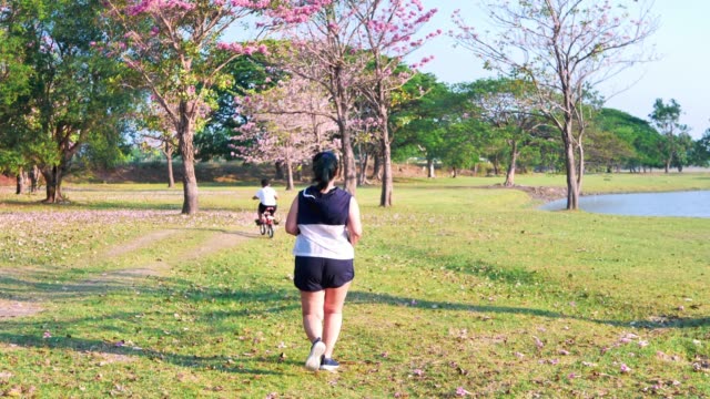 Eine-dicke-asiatische-Frau-joggt-abends-in-natürlichem-Sonnenlicht,-zusammen-mit-seinem-Sohn,-der-mit-dem-Fahrrad-unterwegs-ist.--Für-gute-Gesundheit.-Zeitlupe