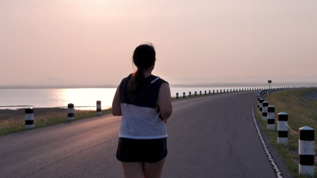 Übergewichtige-asiatische-Frauen-joggen-am-frühen-Morgen-im-Sonnenlicht-auf-der-Straße.-Konzept-des-Gewichtsverlustes-mit-Bewegung-für-die-Gesundheit.-Blick-nach-dem-Blick