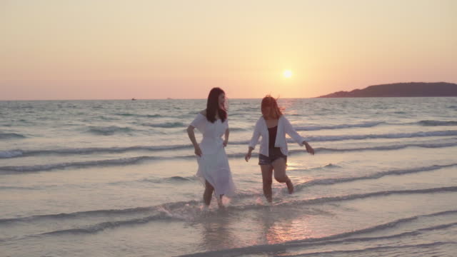 Slow-motion-junges-asiatisches-lesbisches-Paar-läuft-am-Strand.-Schöne-Freundinnen-freuen-sich,-sich-bei-Sonnenuntergang-am-Abend-am-Strand-am-Meer-zu-vergnügen.-Lifestyle-lesbisches-Ehepaar-reist-nach-Strandkonzept