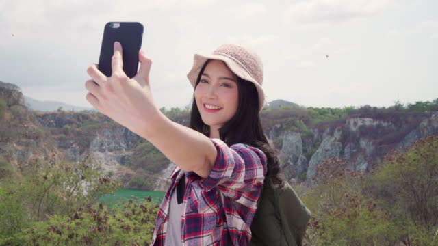 Asian-backpacker-woman-selfie-on-top-of-mountain,-young-female-happy-using-mobile-phone-taking-selfie-enjoy-holidays-on-hiking-adventure.-Lifestyle-women-travel-and-relax-concept.