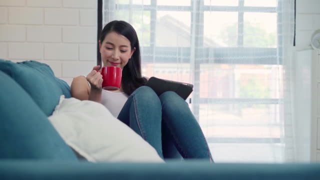Schöne-attraktive-lächelnde-asiatische-Frau-mit-Tablet-mit-einer-warmen-Tasse-Kaffee-oder-Tee,-während-auf-dem-Sofa-liegen,-wenn-Sie-im-Wohnzimmer-zu-Hause-entspannen.-Lifestyle-Frauen-zu-Hause-Konzept.