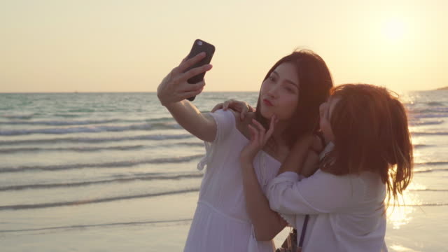 Young-Asian-lesbian-couple-using-smartphone-taking-selfie-near-beach.-Beautiful-women-lgbt-couple-happy-relax-enjoy-love-moment-when-sunset-in-evening.-Lifestyle-lesbian-couple-travel-on-beach-concept