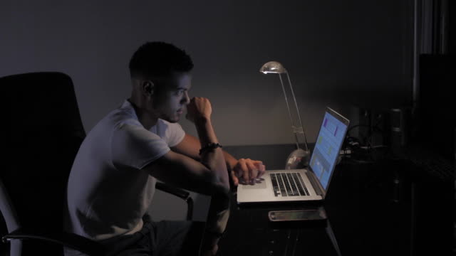 Joven-afroamericano-trabajando-en-computadora-con-lámpara-de-noche-en-su-oficina-oscura-y-negra.-Desplazarse-por-documentos-de-trabajo-y-comprobar-los-mensajes-en-el-teléfono-inteligente.